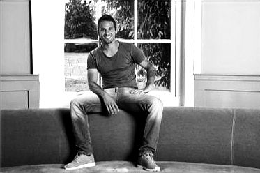 Man in a gray shirt and jeans sits on a brown sofa, smiling. Bright window and greenery in the background create a relaxed mood.