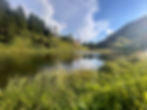 Kleiner Bergsee hinter dem Haus Wallis, mit einem Haus auf einem Hügel im Hintergrund unter blauem Himmel