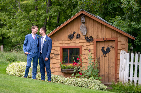Two grooms in blue suits smiling beside a rustic chicken coop.