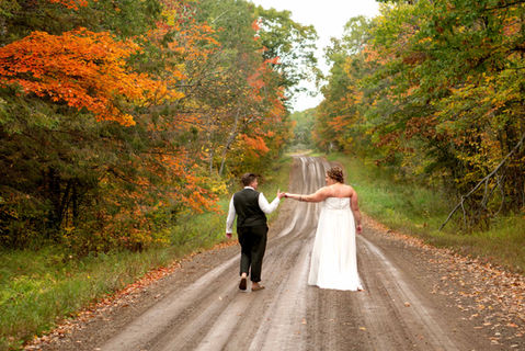 Newlyweds walk hand-in-hand down autumn dirt road, colorful fall leaves.