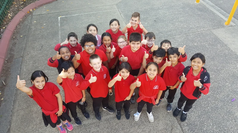 A group of about 15 students in red school uniforms, posing with smiles and thumbs up in a school yard.
