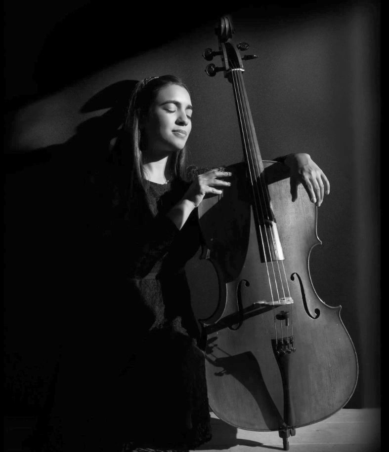 Cellist stands in soft spotlight, eyes closed, holding her instrument against a dark background.