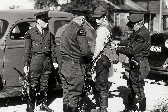 Four uniformed officers frisk a young man beside a vintage car on a rural road in a black-and-white scene. michigan wildlife council protecting michigan's legacy of conservation the tradition is the trophy hunting fishing trapping dnr michigan detroit outdoors