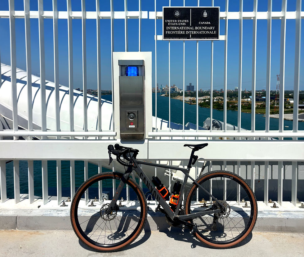 Road bike parked by a white fence at the U.S.-Canada international boundary, with river and city skyline beyond the sign. crossing the gordie howe international bridge multi-use path biking cycling walking running jogging border guide how to detroit michigan windsor ontario canada usa