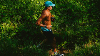 Man in a blue shirt and red vest runs through a forest, holding trekking poles. Background is lush green trees. Mood is energetic. expedition detroit huron 100 register for a 2027 ultramarathon near detroit trail running race adventure endurance athleticism Michigan state parks recreation areas metroparks