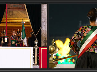La presidenta Claudia Sheinbaum Pardo de espaldas ondeando la Bandera Nacional, luciente de un traje negro con bordados florales multicolores y la banda presidencial desde el balcón central de Palacio Nacional.