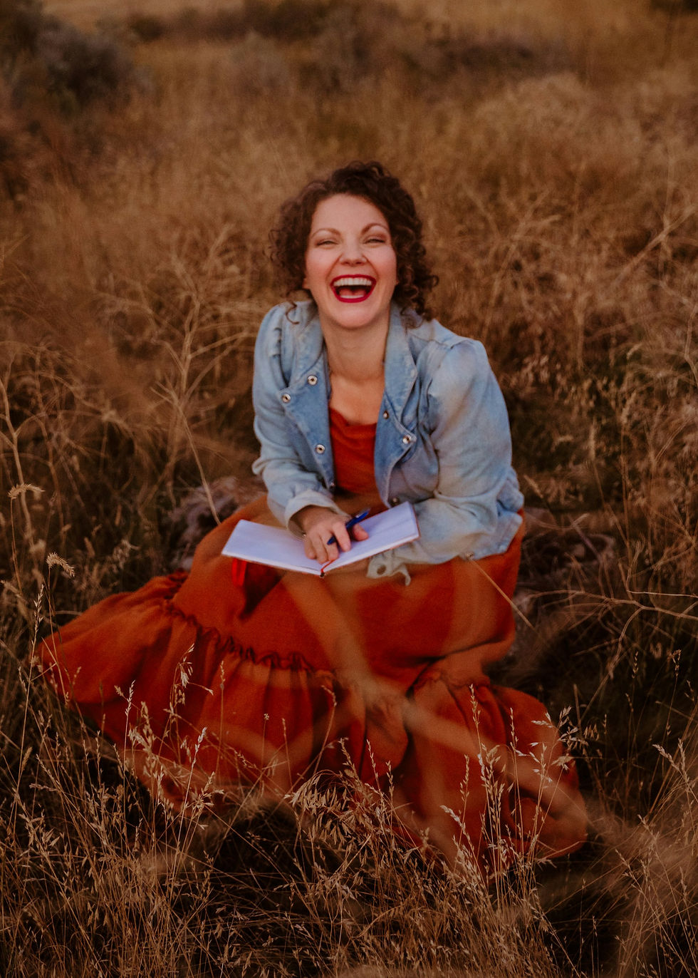 A woman sits in a golden field of tall grass, smiling broadly and laughing joyfully. She is wearing a flowing orange dress paired with a light blue denim jacket. She holds an open notebook and pen in her hands, appearing to be in the midst of writing. The warm tones of the field and her outfit create a cosy and lively atmosphere.