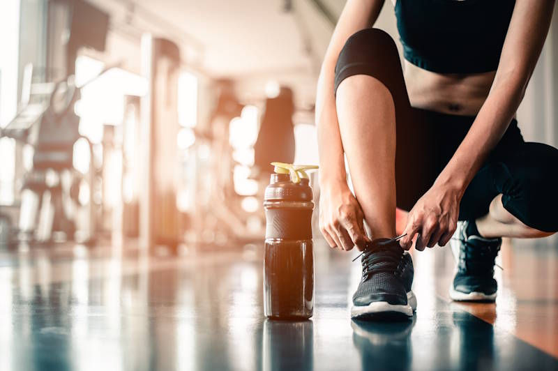 Person tying shoelaces in a gym, next to a water bottle. Bright, modern setting with equipment in soft focus background.