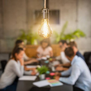 An out of focus photo of a team around a table in an office. With a lightbulb in focus in the foreground.