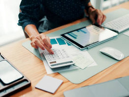A woman sitting at a desk using a calculator and tablet to calculate costs.