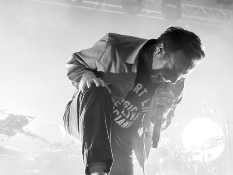 Black and white photo of Sam Carter of the band Architects performing on stage. His right foot sits on a metal riser while he leans into the mic in his left hand.