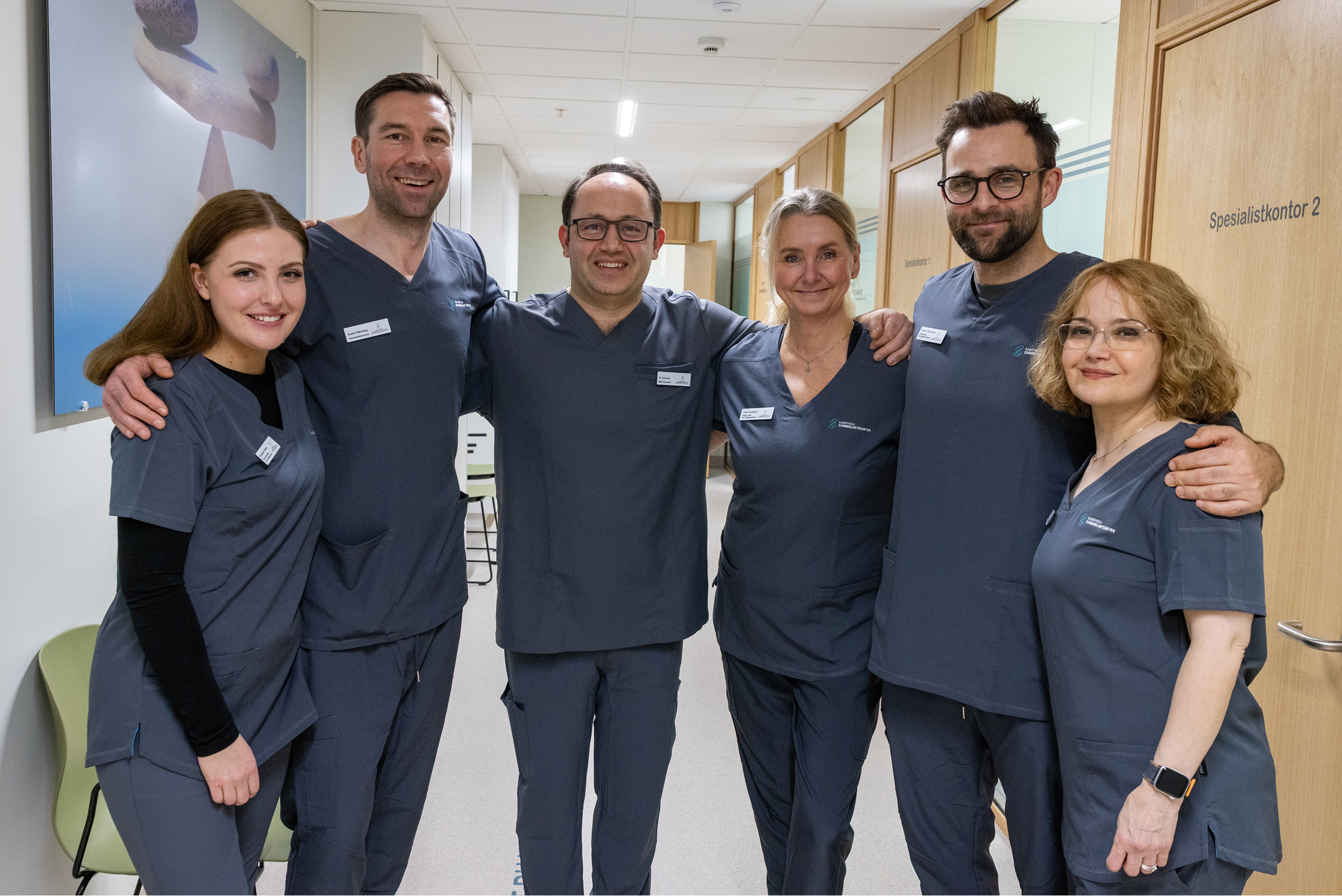 Seks medisinske fagfolk i uniformer hos sandvika svimmelhetssenter smilende i gangen.
