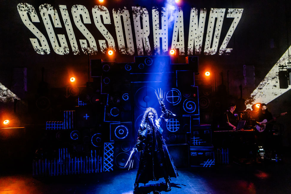 Costume design by Abby Clarke - a leather-clad figure with scissorhands stands spotlit on stage by a microphone