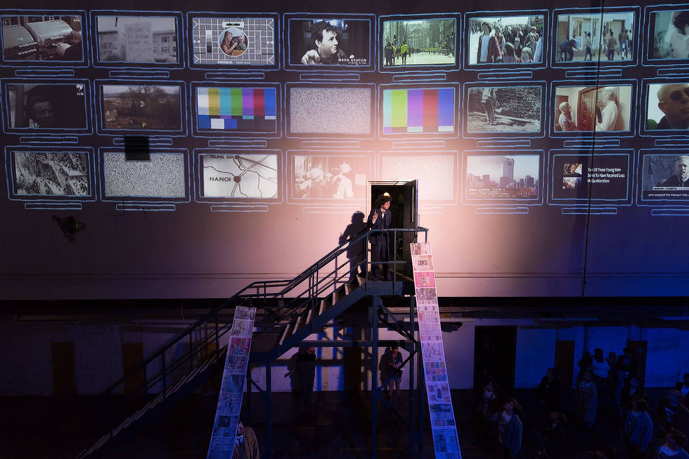 Abby Clarke's set and costume design for City Final, a man stands at the top of a metal staircase, screens are projected behind him and lengths of newspaper reach up to him. 