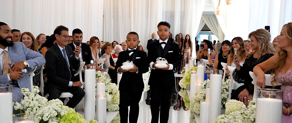 Ring bearers walking down a candle-lit aisle with white florals and sheer drapes during a modern indoor ceremony — Toronto wedding videography.