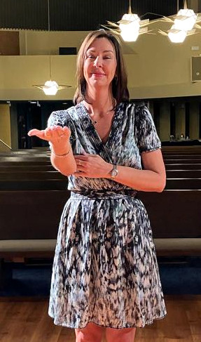 Woman with shoulder-length brown hair, wearing a blue and white dress, stands in front of audience interpreting in ASL.