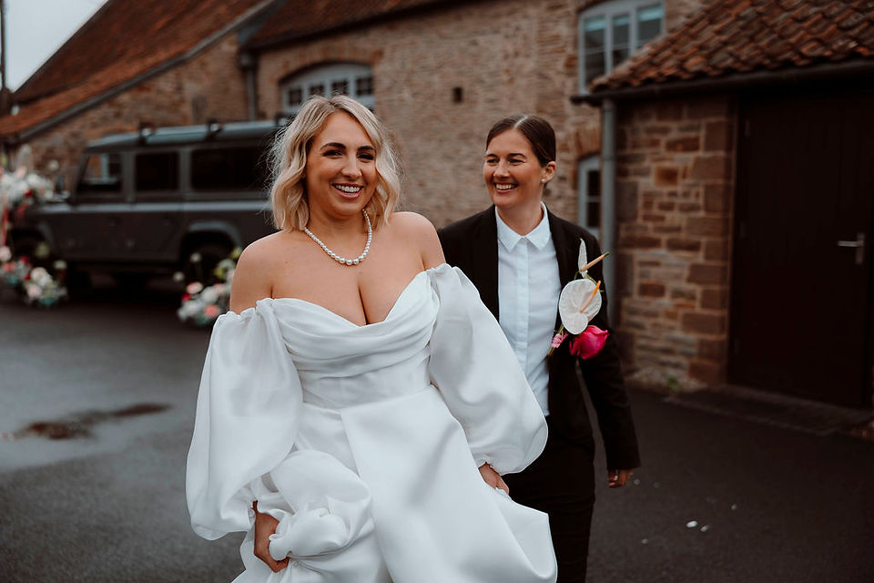 A bride in a satin dress walking in front of bride in a black suit outside with a barn building in the background