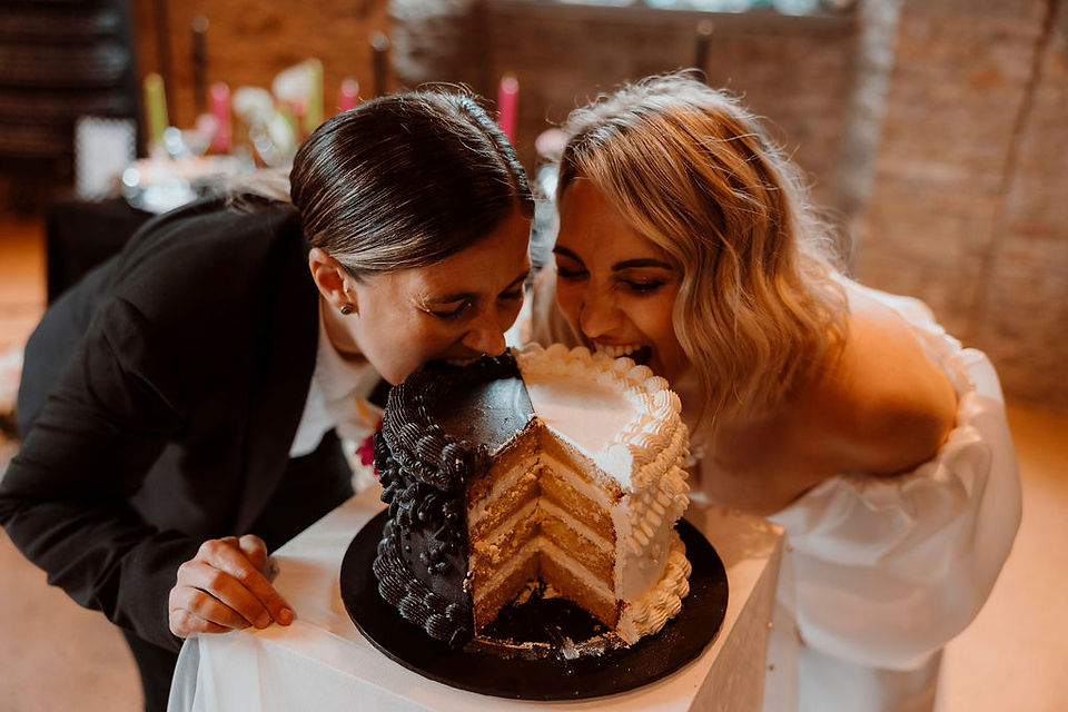 Wedding LGBTQI+ couple leaning down and eating a black and white cake with a slice cut out of it
