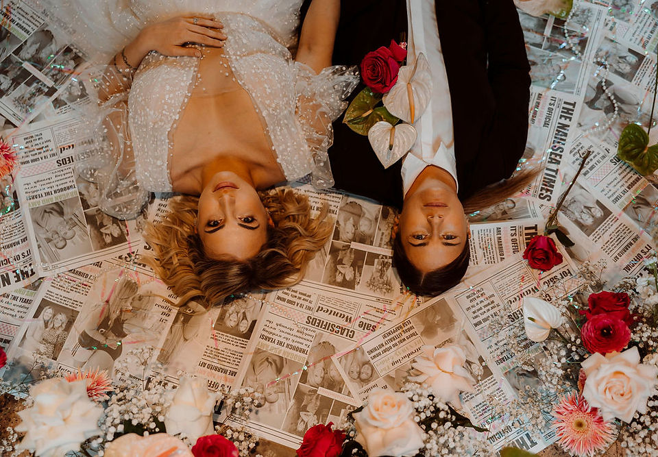 Two brides laying upside down on a wedding newspaper backdrop with pink and red floral displays around the edge