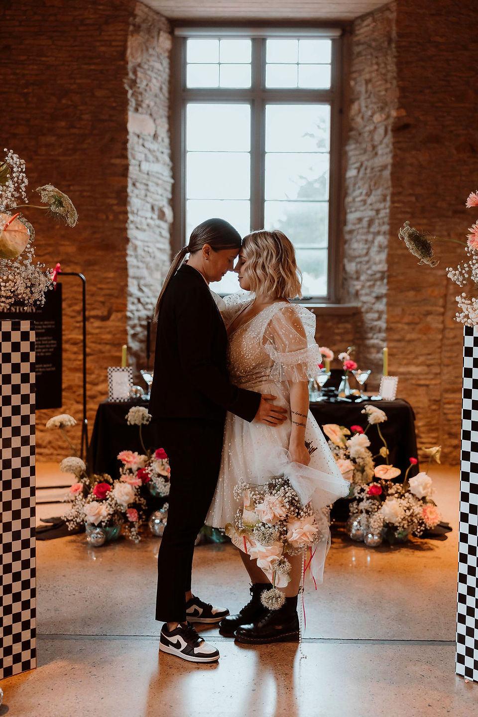 Two brides facing each other in an embrace with their foreheads touching, with floral and checkerboard plinth decor in the background