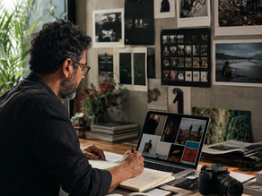 Fotógrafo em um estúdio contemporâneo analisa imagens no notebook, cercado por fotografias impressas, câmera, fotolivro e anotações, em uma cena que representa curadoria, leitura de mercado e reflexão sobre os rumos da fotografia profissional.
