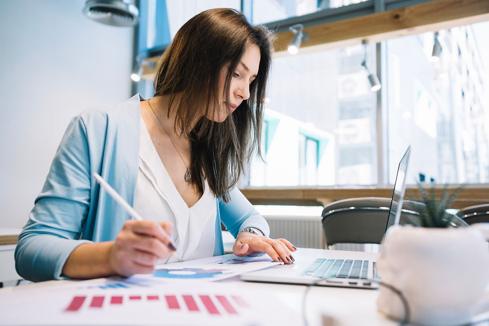 Woman in a bright office writes on charts beside a laptop, focused on work; blue cardigan and red graphs visible.