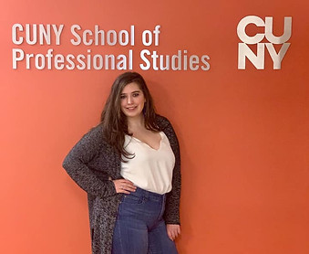 Woman with dark hair, wearing jeans and white shirt standing in front of orange wall with wording "CUNY School of Professional Studies"