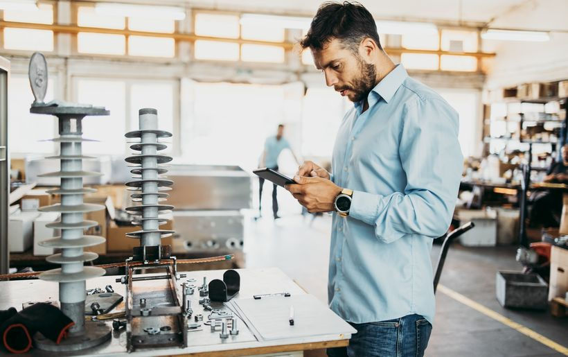 Homem em ambiente industrial analisa equipamentos sobre uma bancada enquanto utiliza um tablet, com máquinas e peças ao fundo.