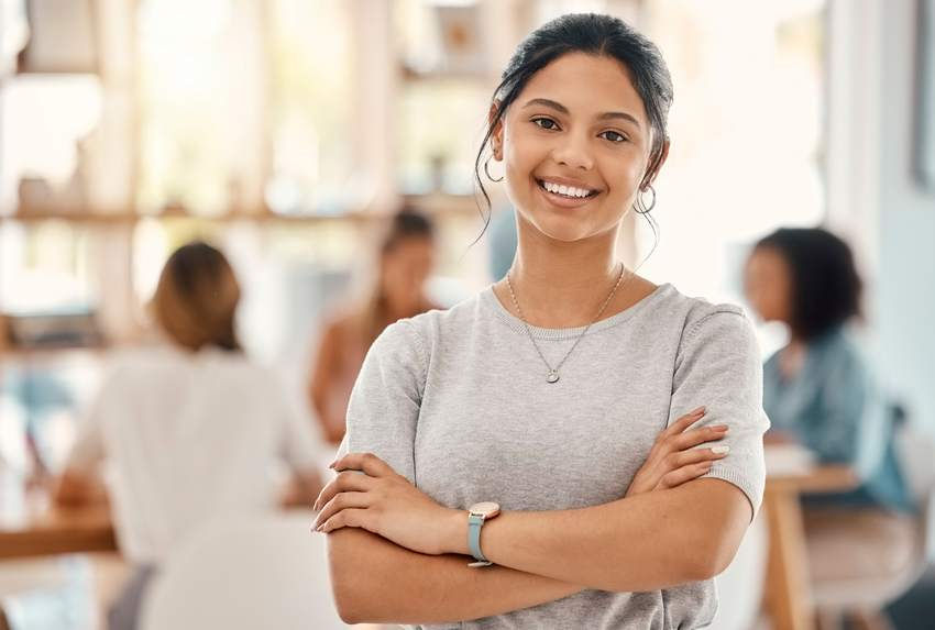 Jovem mulher sorri confiante de braços cruzados em ambiente moderno de escritório, representando o mercado de trabalho e oportunidades de estágio.