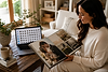 Custom photo album design service showing a woman reviewing a finished heirloom album while digital photographs remain on a laptop, representing the journey from digital files to professionally crafted albums and wall art.