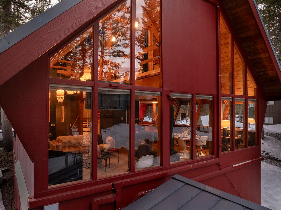 Red A-frame cabin with tall windows glowing warm inside, set among snowy trees at dusk.