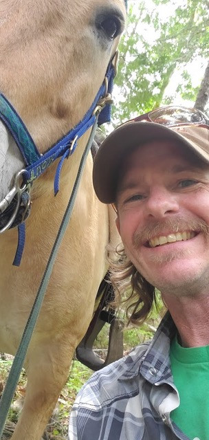 Smiling person with mustache and cap standing next to horse with trees in background