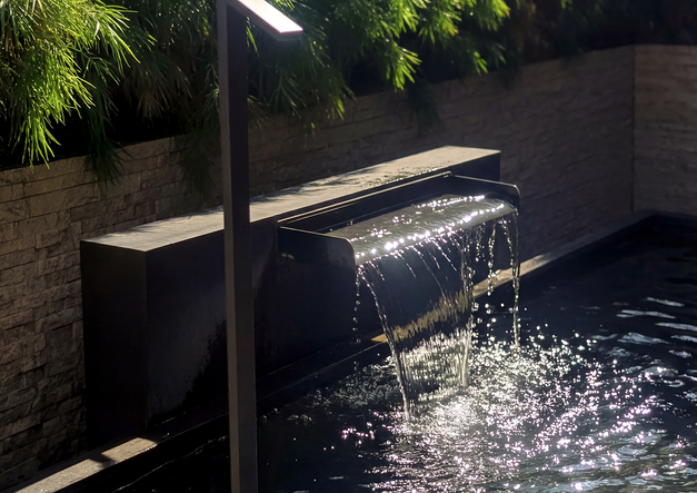 Close-up of a sleek, modern, dark metal waterfall fountain with lush garden backdrop, designed for a luxury Los Altos, CA landscape.