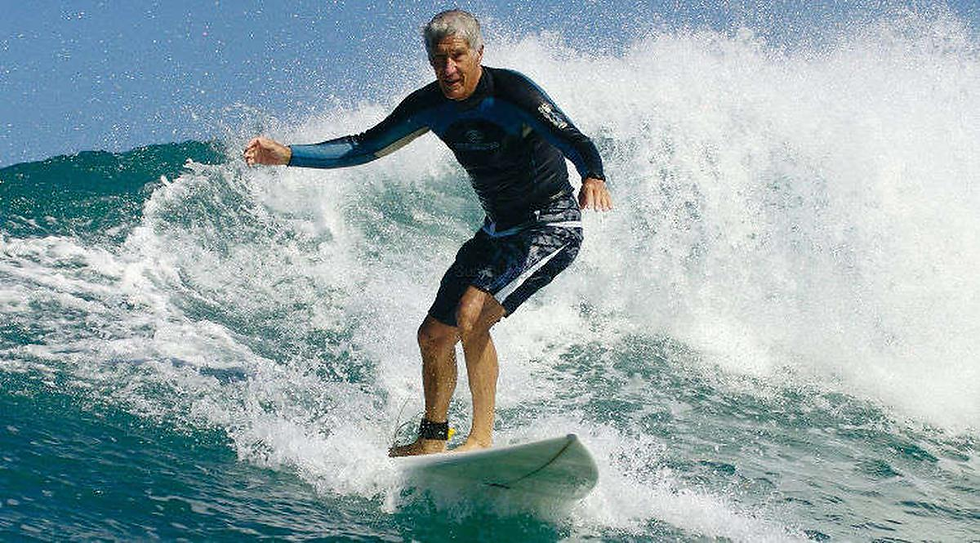 Surfeur âgé (Joël de Rosnay) en combinaison noire, en équilibre sur une vague turquoise sous un ciel bleu, avec éclaboussures blanches.