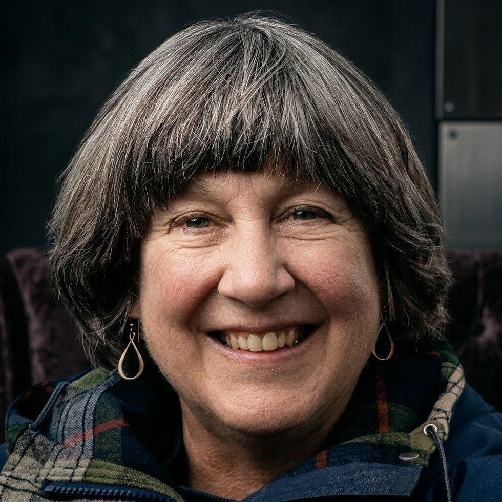 Portrait d’une femme souriante (Donella Meadows) aux cheveux gris en carré, boucles d’oreilles pendantes et écharpe à carreaux, fond sombre.