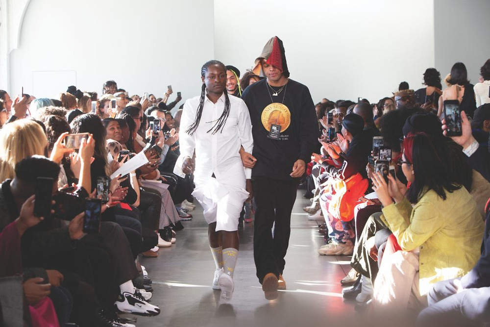 A diverse group of models walk down a runway, flanked by spectators
