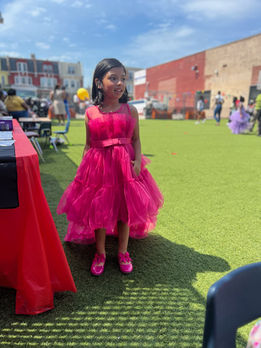  A little girl in a pink prom dress happily stands on a green grassy field, enjoying a sunny day outdoors.