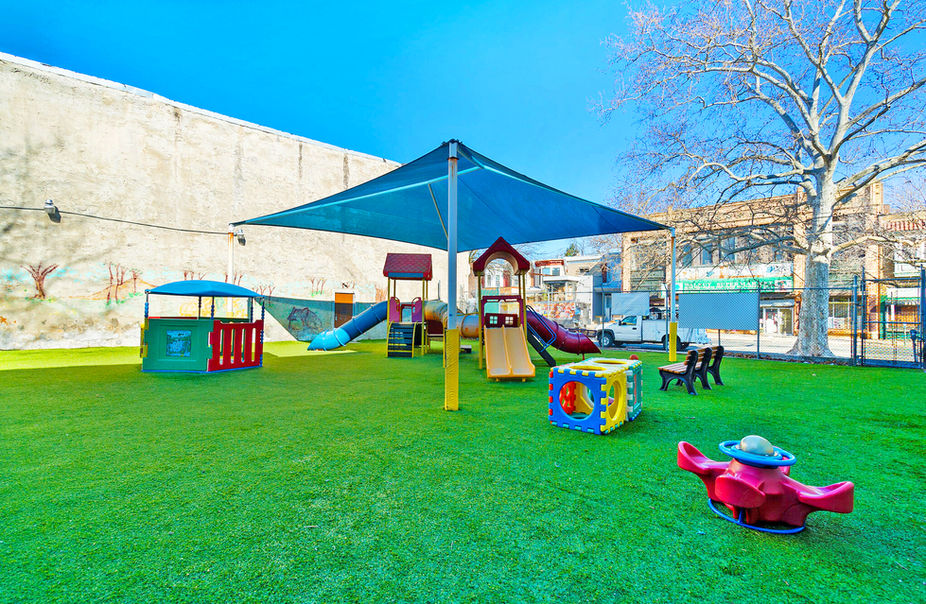 A spacious outdoor play area with climbing equipment, slides, and a shaded canopy. The artificial turf provides a safe and fun environment for preschool children.