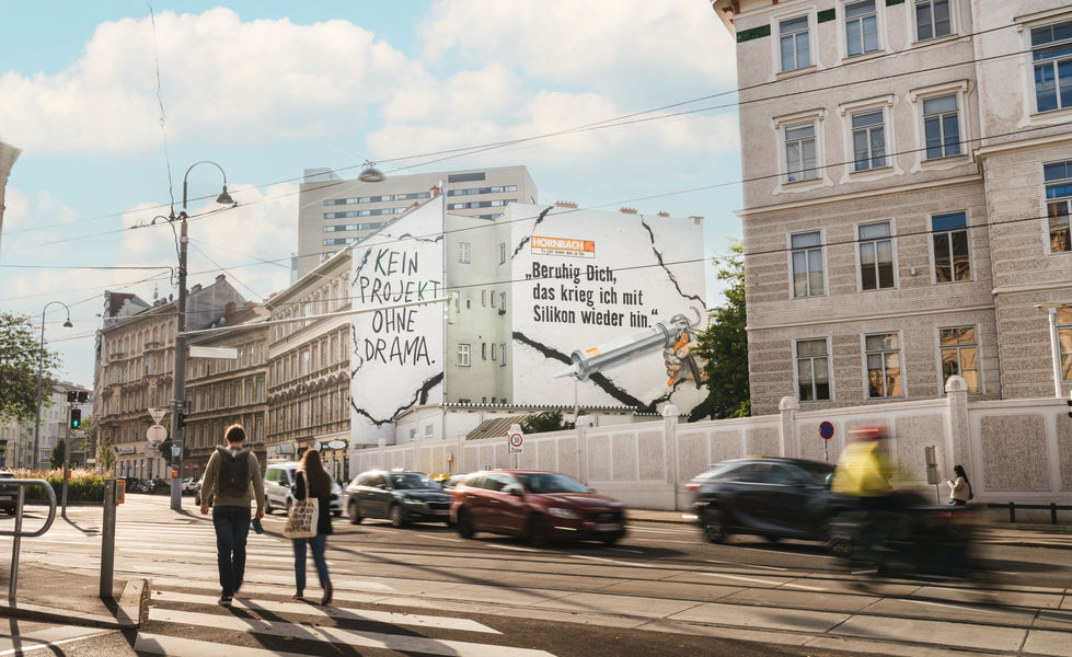Die Hornbach-Wandbemalung aus der Ferne gesehen an einer Hausfassade in Wien. Im Vordergrund eine belebte Straßenkreuzung mit fahrenden Autos und vorbeigehenden Passanten, alles in Bewegung.