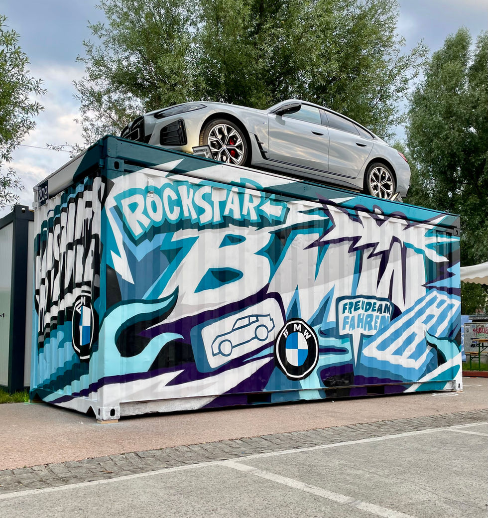ein bemalter Container mit Graffit bemalt, Graffiti Werbung für BMW, Darauf ein BMW Auto, blaue, weiße und türkis Elemente, dahinter Bäume, davor ein Parkplatz, Linz