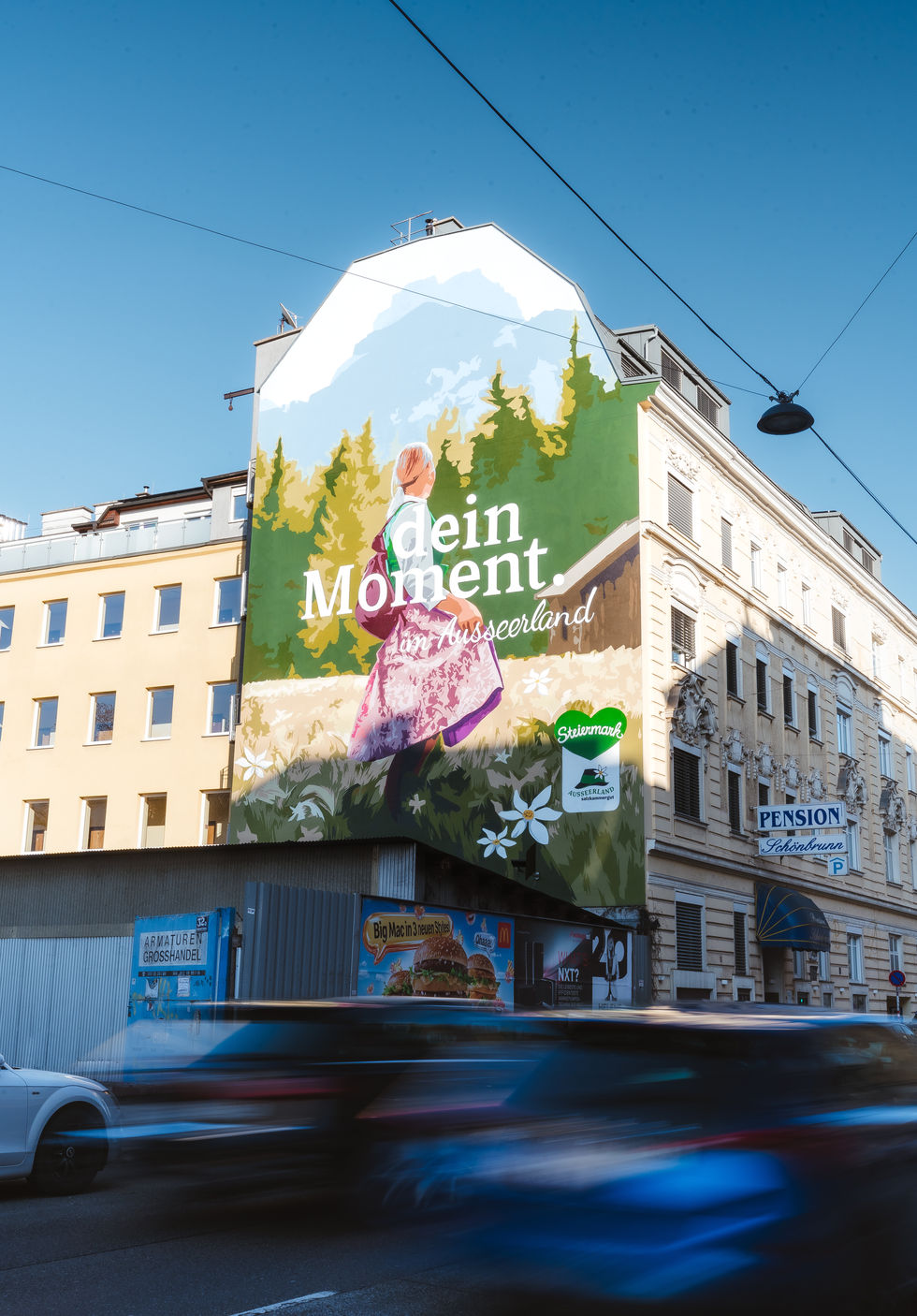 Großformatiges Wandgemälde an einer Gebäudefassade an einer stark befahrenen Straße: Eine Frau im Dirndl steht auf einer Wiese und blickt auf eine stilisierte Berg- und Waldlandschaft vor blauem Himmel; darüber der Schriftzug „dein Moment. im Ausseerland“.