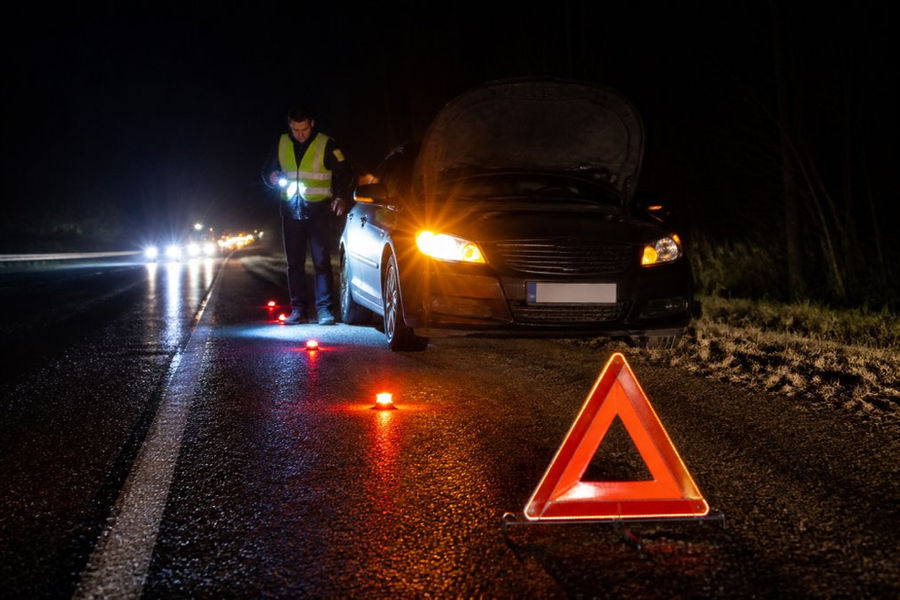 Nighttime scene of a disabled vehicle with headlights and hazard lights illuminated on the highway shoulder, with flares and reflective triangles glowing around it, a driver in a safety vest visible by flashlight