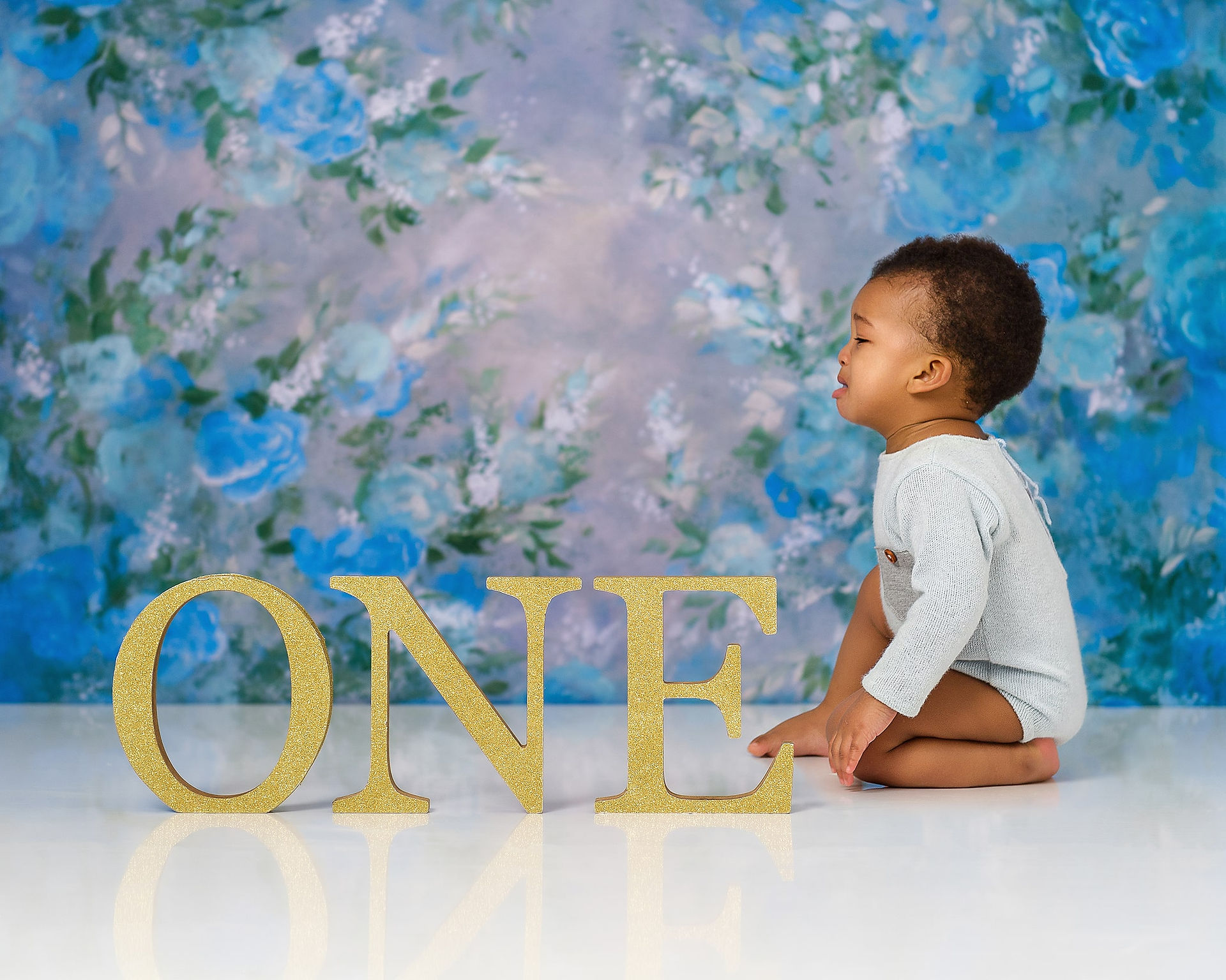 Boy wearing a blue romper is looking at one golden wooden letters during his first birthday photoshoot