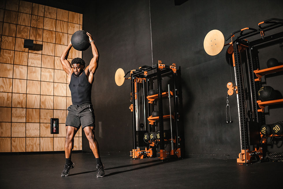 Man exercising in a gym, lifting a black medicine ball overhead. Wooden panels and gym equipment in the background. Determined mood.