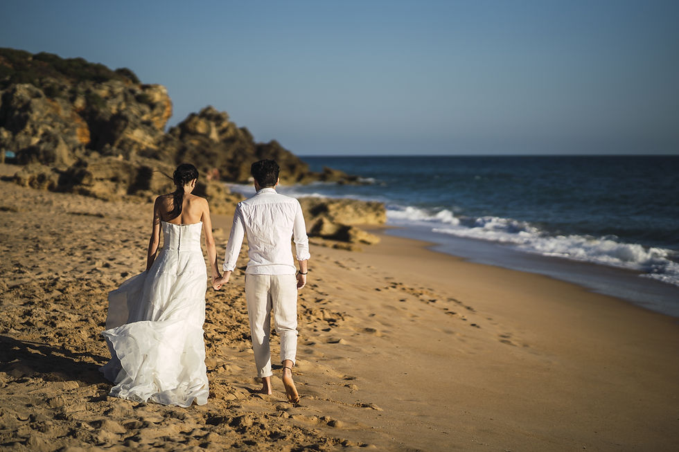 Wedding in Curaçao, A couple in white attire walks barefoot on a sandy beach by the ocean with rocky cliffs under a clear blue sky.