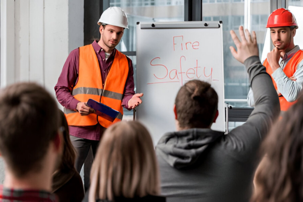 Two men in safety vests and helmets lead a fire safety training. One man writes on a board. Attentive audience, visible raised hand.