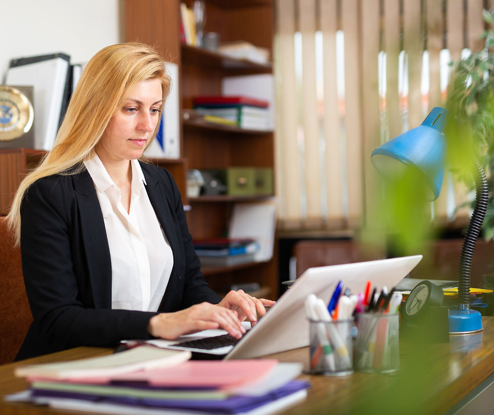Woman in a black blazer types on a laptop at a cluttered desk. Blue lamp, office shelves, and blinds create a focused work setting.