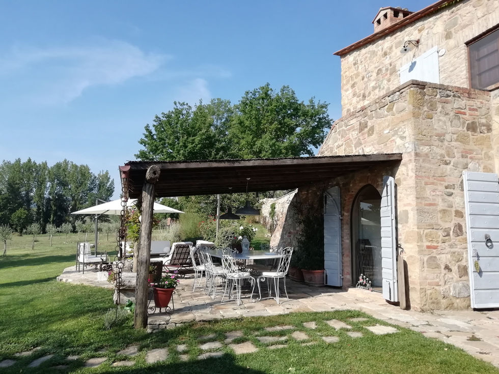 Covered timber pergola terrace at Podere Morone Montepulciano with wrought-iron furniture, stone farmhouse wall, and open Tuscan countryside garden