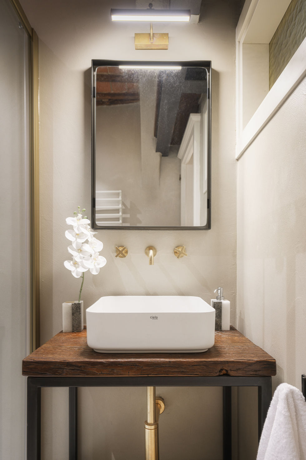 Bathroom vanity with white vessel sink, brass wall-mounted faucet, dark-framed mirror, and white orchid in Appartamento Montepulciano, Tuscany