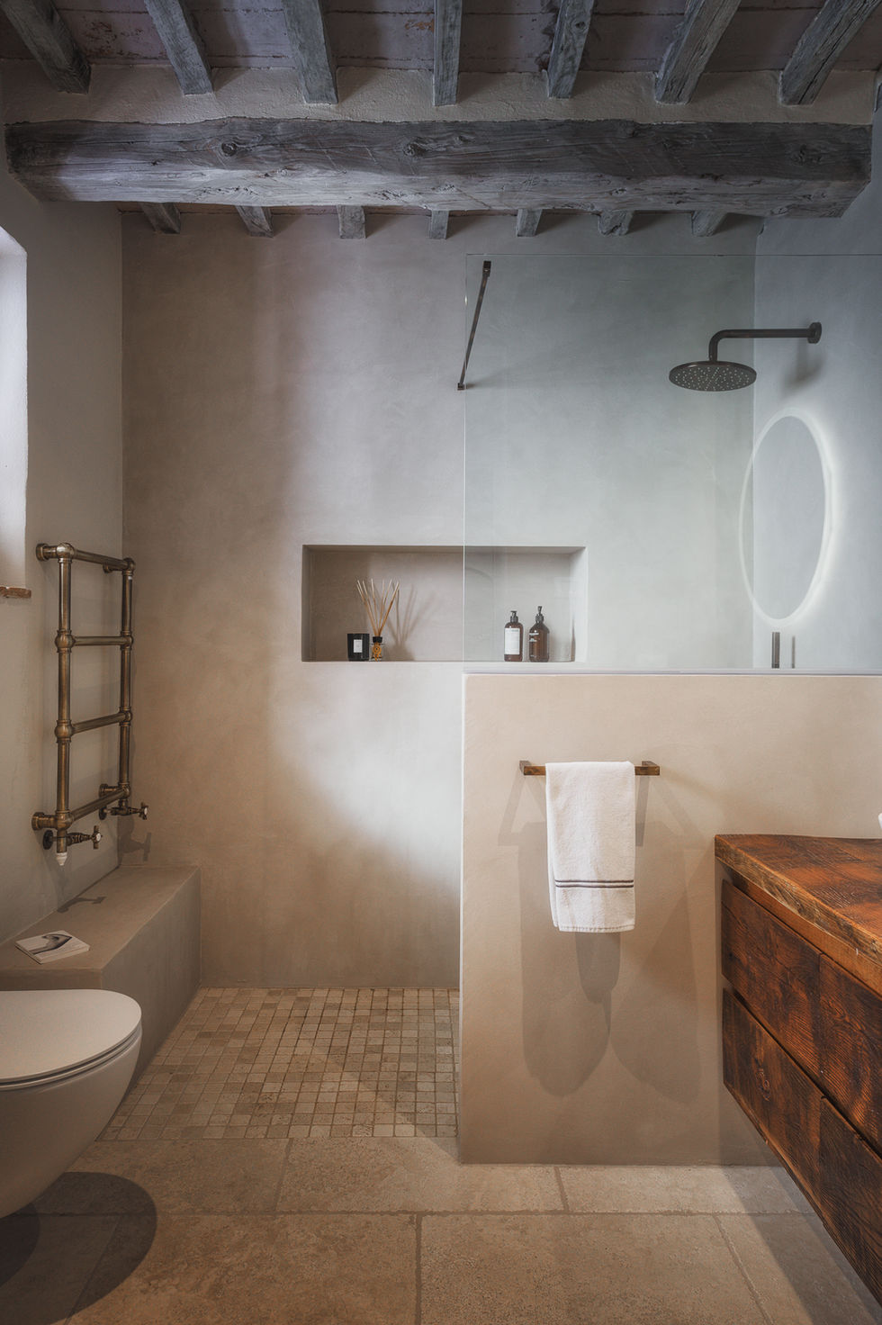 Bathroom at Podere Madonna, Montefollonico showing microcement shower partition, built-in niche, brass towel rail, reclaimed wood vanity, and oval backlit mirror